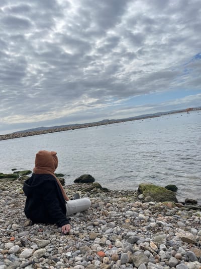 The image displays a single individual seated on a rocky beach in L'estartit, Spain, facing a body of water. The subject is viewed from behind, wearing a dark hooded garment, light-colored trousers, and a distinct brown, textured hood with ear-like protrusions, paired with a brown scarf. The left hand of the individual rests on the pebbles. The immediate foreground consists of a densely packed surface of grey, brown, and white pebbles and small rocks. Larger rocks at the water's edge are covered with green marine growth.

The midground features calm, rippled grey-blue water, extending towards a distant horizon. A long, low rock breakwater or jetty extends from the left side of the frame into the distance, separating the inner bay from the open water. Multiple small yellow buoys are visible in the water beyond the breakwater, and a small, light-colored navigational marker or structure stands on the breakwater further along. In the background, low-lying hills or landmasses are visible along the distant horizon. The sky is predominantly overcast with dense, grey clouds, featuring lighter areas and glimpses of faint blue.