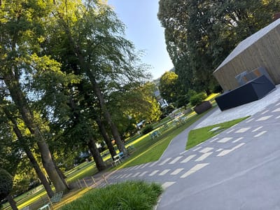 This media file depicts a park-like setting with a modern building, possibly a restaurant or event space, on the right. The building has a natural wood exterior and a sloped roof. In front of it, several large, dark, covered objects are placed, which might be outdoor furniture or equipment. A paved pathway with a geometric pattern of light and dark gray stones leads through the scene.

The park itself is well-maintained with lush green grass and tall, mature trees casting long shadows, indicating the late afternoon or early evening. Several groups of empty chairs and tables, some made of metal and plastic in blue and green, are scattered across the lawn, suggesting outdoor seating areas. A few people are visible in the background, strolling or sitting on the grass, adding a sense of calm and leisure to the scene. One person is walking a dog on the grass. The sky is clear and blue, hinting at pleasant weather. In the distance, more trees and some buildings, possibly houses, are visible, with a gentle hillside in the far background. The overall atmosphere is peaceful and inviting, suggesting a relaxed day in a scenic outdoor location. There is no visible text in the image.