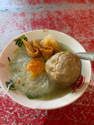 A close-up, overhead shot captures a bowl of bakso, a popular Indonesian meatball soup, served on a red, distressed table. The bowl is white with a faint green and black logo on the rim, partially obscured by the food. Inside, a large, single meatball, roughly the size of a fist, is the dominant feature. It's a light brown, textured sphere.
Beneath the meatball, a bed of clear, thin noodles, likely bihun or soun, is visible in a light broth. To the left of the meatball, a dollop of vibrant orange chili paste sits atop the noodles. Three fried wonton wrappers, golden brown and folded into decorative shapes, are positioned above the noodles and to the left of the meatball. Scattered throughout the broth are small, chopped green herbs, likely scallions or cilantro, adding a touch of freshness. A silver spoon rests on the right edge of the bowl, its handle extending out of frame. The background is the red table, characterized by its worn, crackled surface with patches of white and darker, almost black, accents, suggesting age and heavy use. The setting is likely a local eatery in Sungai Sipai, Indonesia, given the context of the dish. No people are visible in the image.