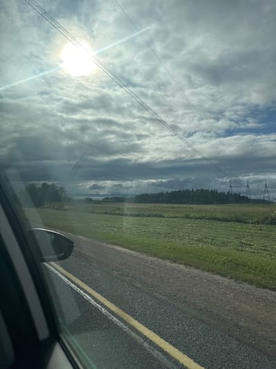 The image captures a view from a vehicle traveling along a paved road with a yellow line dividing the lanes. To the right of the road is a green field. In the distance, a dense line of trees forms a dark horizon against a dramatic sky filled with heavy, grey clouds. The sun is a bright, intense spot in the upper left portion of the sky, casting a glare that illuminates the clouds and creates lens flare effects. Several high-voltage power lines stretch across the sky, supported by distant towers visible on the right side of the image. The overall impression is of a rural landscape under a dynamic sky.