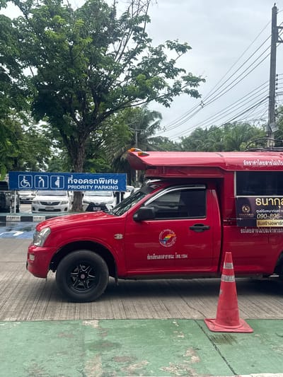 In the city of Mae Hia Town Municipality, Thailand, a red pickup truck, modified into a songthaew (a shared taxi), is parked on a concrete surface. The songthaew has a covered passenger area on the back and is adorned with decals and signage, including Thai script indicating it is a transportation service. Notably, one decal features a phone number: 090-896-3626. Adjacent to the songthaew, a blue banner is suspended, displaying symbols for disabled parking and pregnant women, along with Thai text and the English phrase "PRIORITY PARKING." Several other vehicles, including white cars, are parked in the background. The scene is framed by lush green trees and overhead electrical wires, with an orange traffic cone placed on the ground in the foreground, suggesting a traffic management or parking area. The sky is overcast.