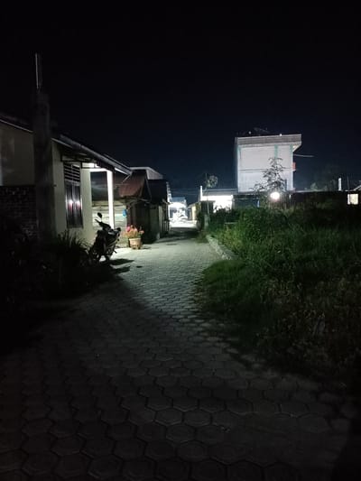 The image captures a nighttime scene in Bireuenmeunasahreuleut, Indonesia. The setting appears to be a pathway or alleyway between buildings, illuminated by artificial light sources, suggesting it is after sunset.
The foreground is dominated by a paved pathway made of hexagonal interlocking stones, which recedes into the distance. To the left, a section of a building is visible, with a window reflecting light and a motorcycle parked in front of it. Beside the motorcycle, a planter holds pink flowers. Further down the path, more buildings are visible, with some showing signs of corrugated metal roofing. On the right side of the path, overgrown grass and vegetation create a dense, dark border. In the background, a taller, multi-story building stands out, its facade illuminated.
The time of day is clearly night due to the dark sky and the artificial lighting. The weather is not explicitly indicated, but the dark sky and the need for artificial light suggest it is not daytime. The location cues, such as the architecture of the buildings and the type of vegetation, are consistent with a tropical or subtropical region, aligning with the provided location context of Indonesia.
No specific activity or event is evident; the scene appears to be a still, everyday view of the area at night. The overall mood is quiet and perhaps a little somber due to the darkness and the dim lighting. There is no visible text in the image.