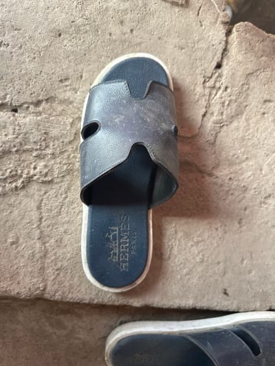 A single, dark blue Hermès slide sandal rests on a rough, grey concrete surface. The sandal features the iconic "H" logo embossed on the top strap. The insole of the sandal displays the Hermès logo and "PARIS" in white lettering. The concrete surface appears weathered and textured, with visible cracks and imperfections. The lighting suggests an outdoor setting. The image is a close-up, focusing on the sandal and its immediate surroundings. No people are visible in the image. The location in the city of International, Kyrgyzstan is unknown.