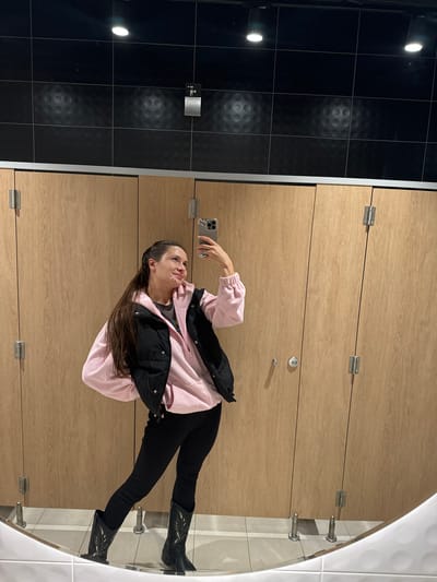 A woman is taking a selfie in a restroom. She is wearing black pants and cowboy boots, a pink hooded sweatshirt, and a black puffer vest. The restroom has light-colored wooden stall doors, black tiled walls, and a mirror. It is unknown whether the location is in Ivashevo, Russia.