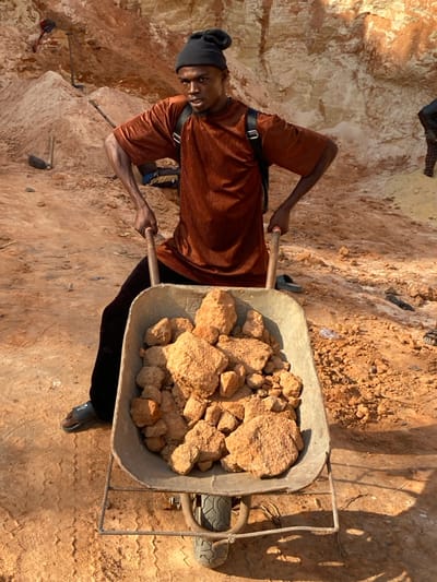 A person stands next to a wheelbarrow filled with dirt and rocks, possibly in a mining or quarry setting. The individual wears a brown shirt and a black hat. They appear to be holding the handles of the wheelbarrow, ready to move the load. The background shows a dirt wall or hill, suggesting an excavated area. A few tools such as a shovel are visible nearby.