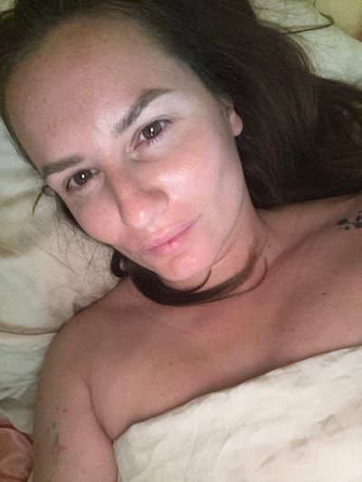 A close-up, overhead selfie shows a woman lying in bed, with her face filling most of the frame. Her head is turned to the left, and her eyes are open and looking at the camera. She has long, dark brown hair that is spread out around her head. Her skin appears to be smooth and clear, with a light dusting of freckles across her nose and cheeks. She is wearing a light-colored top that is partially visible, and she is covered by a white blanket. The background is a soft, blurry beige, suggesting a bedroom environment. The image does not contain any discernible information about the location in the city of Буран Снт, Russia.