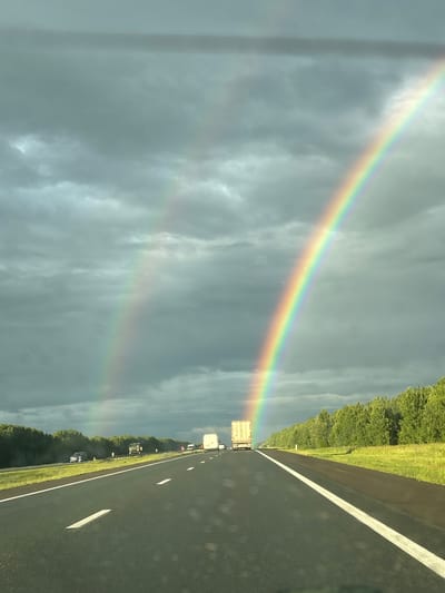 The image captures a scenic view from a moving vehicle on a highway in Shymkovo, Russia. The primary subjects are a double rainbow arcing across a cloudy sky and the highway itself, lined with lush green trees and grass. Several vehicles, including trucks and cars, are visible on the highway, suggesting ongoing travel. The weather appears to be overcast with the presence of rain, as indicated by the faint water droplets on the camera lens and the dramatic cloud formations. The time of day is likely late afternoon or early evening, given the subdued lighting and the arc of the rainbows. The overall mood is serene and majestic, enhanced by the natural phenomenon of the double rainbow against the moody sky. No visible text is present in the image.