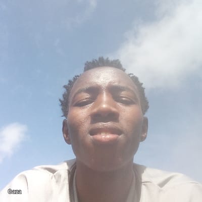 The image depicts a close-up, low-angle selfie of a male individual, appearing to be of African ethnicity, under a clear blue sky with scattered white clouds. The subject has short, dark, curly hair and an even skin tone, with visible light reflection on the forehead and nose, suggesting a shiny or slightly sweaty complexion. His eyes are open, looking directly at the camera, and his mouth is slightly open, revealing teeth. He is wearing a light-colored, possibly beige or light grey, short-sleeved top over a darker undershirt. The lighting is bright, indicating outdoor daytime conditions. The word "Gaza" is present in white text in the bottom-left corner of the image. The scene is located in Bonyamaro ward, Kenya.