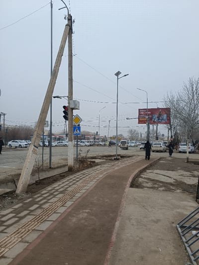 An urban scene depicts a street with sidewalks and a crosswalk, featuring a tilted utility pole supported by another. Pedestrian crossing signs, traffic lights, and streetlights are visible. The street contains vehicles and people. A billboard is in the background. The day appears overcast. The sidewalk has a tactile paving surface.