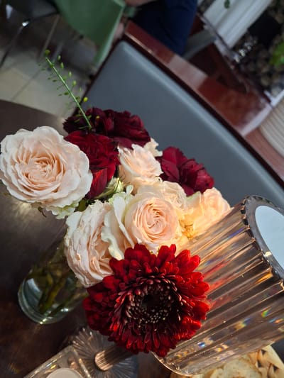This image features a close-up shot of a flower arrangement in a glass vase, set on a wooden table. The bouquet consists of several cream-colored roses, dark red carnations, and a prominent deep red gerbera daisy. The flowers are artfully arranged, with some roses in full bloom and others still opening. A decorative glass object, possibly a candle holder or vase, is positioned to the right of the flower arrangement, catching and refracting light. The background is softly blurred, suggesting an indoor setting. Hints of furniture, like a dark chair with a padded seat and a wooden surface with a reddish hue, can be discerned. The lighting appears to be artificial, casting a warm glow on the flowers and creating highlights on the glass. The overall mood is elegant and serene. No people are clearly visible, though a sliver of dark blue fabric, possibly clothing, is present in the far left background, indicating the presence of someone out of frame. No text is visible in the image. The context of Pila, Poland, does not provide specific visual cues within the image itself.