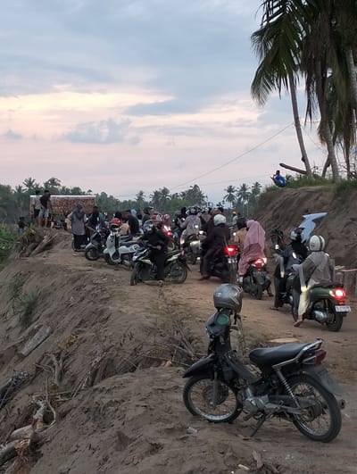 The image depicts a dirt road elevated on an embankment in what appears to be Kapa, Indonesia, evidenced by the license plates on some of the motorcycles and the tropical vegetation. A large group of people on motorcycles is gathered on the road. Some are parked, while others appear to be waiting or moving slowly. In the foreground, a black motorcycle is parked with a helmet resting on its seat. The embankment is made of soil and has exposed roots and debris visible. In the background, palm trees and other vegetation are visible, along with a small structure with a thatched roof. The sky is overcast, suggesting either early morning or late afternoon. One person is sitting on top of a tree. Several people can be seen standing near the small thatched roof.