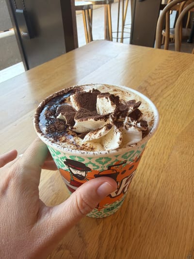 A person's hand holds a takeaway cup filled with a whipped cream-topped beverage, likely a coffee or dessert drink. The cup has a colorful design featuring cartoonish figures and geometric patterns. Generous swirls of whipped cream are visible at the top of the drink, dusted with a dark powder, possibly cocoa or cinnamon. The beverage is in a paper cup, typical for to-go orders. The setting appears to be a cafe or a casual dining area, with a wooden table surface in the foreground and background. The lighting suggests it might be daytime, possibly late morning or afternoon, with natural light falling on the scene. In the background, elements of seating, possibly chairs with curved wooden backs, are partially visible, indicating an indoor or covered outdoor space. A dark rectangular object with some text on it is present in the background, on a dark surface. The overall impression is a snapshot of a person enjoying a drink in a relaxed setting. The image is oriented in a way that the cup is tilted, with the hand gripping it from the side.