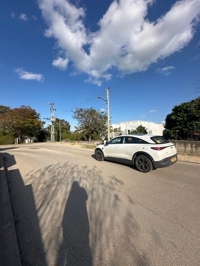 The image depicts an outdoor street scene in Beit Yitshak Sha'ar Hefer, Israel, under a bright blue sky with scattered white cumulus clouds. A white crossover SUV, featuring black lower body trim and dark wheels, is positioned on the right side of an asphalt road, facing away from the viewer. The vehicle displays an Israeli license plate reading "718:9D". A prominent shadow of a person, consistent with the photographer, extends from the lower left foreground across the cracked asphalt surface of the road. On the left side of the road, a tall metal utility pole equipped with electrical components and crossarms stands near a cluster of trees. On the right, a wooden utility pole with a streetlamp and overhead power lines is visible. In the background on the right, a light-colored, low-rise building with a flat roof is partially obscured by foliage. Trees line both sides of the street, displaying green and some reddish-brown leaves. The scene is brightly illuminated by direct sunlight, casting distinct shadows.