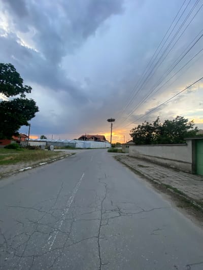 The image captures a view of a cracked asphalt road in Miriantsi, Bulgaria, leading into the distance under a dramatic sky filled with dark clouds tinged with the orange glow of sunset. To the left, a large green tree stands beside a red-tiled roof house, and further along, a row of white-sheeted greenhouses. On the right side of the road, a low, plastered wall with a green gate borders the property. High above, power lines crisscross the sky. In the distance, a utility pole supports a large stork's nest, with a stork visible within it. The overall impression is one of a rural or suburban setting during twilight.