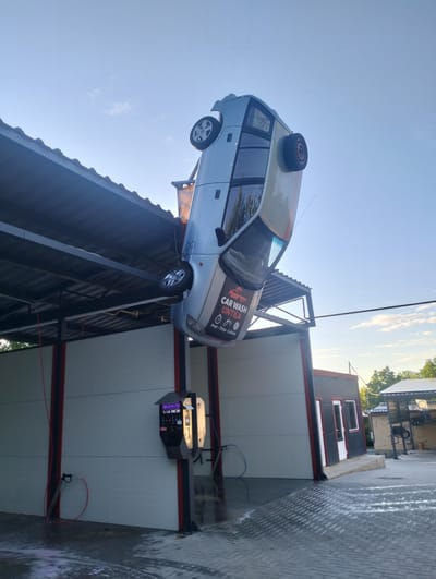 A light-colored car is suspended upside down from the roof of a car wash in Șoldănești, Moldova. The car wash appears to be a self-service facility, with hoses and a payment station visible. The car is positioned at an angle, with its wheels facing the sky. The car wash building is white with red trim. The sky is blue with a few clouds. The ground is paved with bricks. Trees are visible in the background.