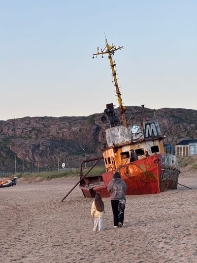 A rusted shipwreck lies beached on a sandy shore in Teriberka, Russia, under a clear sky. The ship, tilted on its side, is adorned with graffiti. Two people, a woman in a grey hoodie and a young girl in a yellow jacket, walk away from the shipwreck across the sand. In the background, rocky hills rise behind a grassy area with streetlights and a fence. Further in the distance, modern buildings with glass facades are visible. Another smaller, partially visible shipwreck lies to the left of the main subject.
