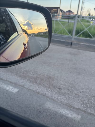A side-view mirror reflects a sunset sky and a road with a car. The mirror shows a dramatic cloud formation illuminated by the setting sun, with a car visible in the distance on the road. The reflection also shows the side of a red car, indicating that the photo was taken from inside a vehicle. The environment outside the mirror appears to be a road with a fence and some greenery. There are no people visible in the reflection or the immediate surroundings. The image was taken in the city of Akberdino, Russia.