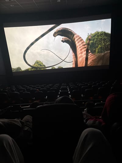 A low-angle, off-center shot inside a movie theater. A large screen displays two long-necked dinosaurs with brown-striped patterns against a blue sky and green trees. Rows of dark theater seats are partially visible on the right, occupied by blurry figures. The person taking the picture is in the foreground in the lower right. The lighting is dim, casting shadows across the theater.