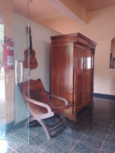 The image shows a building interior in Horqueta, Paraguay. A glass partition separates the foreground from the main interior space. Mounted on the wall behind the glass are a red fire extinguisher and a guitar. A wooden chair rests against the wall, partially obscured by the glass. To the right of the chair is a large, dark wooden wardrobe. The floor is tiled with dark green and gray square tiles. Sunlight is visible through an opening in the glass, indicating an exterior. The building's interior walls are a light, warm color. A decorative element is visible in the distance to the right of the wardrobe. A glass door frame is visible to the left. A light switch is visible on the wall between the fire extinguisher and guitar.
