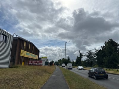 The image presents an overcast daytime scene of a roadside in what appears to be a commercial or industrial area in Reading, United Kingdom. The sky is dominated by large, textured gray and white clouds, with patches of lighter, slightly blue sky visible.
On the left, a modern building with corrugated metal siding is adjacent to a brick building featuring yellow-framed windows and a distinctive curved roofline. This brick building prominently displays large yellow banners advertising "ABBEY WINDOWS," listing services such as "Orangeries," "Conservatories," "Windows - Doors," and "Bifolding Doors." The website "www.abbeywidows.co.uk" and a phone number (0118 9666666) are also visible. Below these, a red sign boldly states, "WE WON'T BE BEATEN ON PRICE," alongside "ReadingTradeWindows.co.uk." Further down the road, another sign for "ABBEY Reading Trade WINDOWS" is visible, featuring brands like "origin," "ALUCO," and "hup!".
A paved sidewalk runs along a dry, grassy verge on the left. To its right, a multi-lane road carries traffic. Vehicles visible include a black hatchback, identified as a Toyota Aygo by its design, with the UK license plate "EX14 BOV" in the foreground. A white car and two white vans are further along the road. Streetlights line the road, and tall evergreen trees form a dense green backdrop along the right side of the street. The scene depicts an active but routine commercial roadside environment.