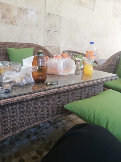 An outdoor scene features a glass-topped rattan table with various items. On the table, there is a large brown plastic jug containing a light brown liquid, a partially full bottle of Fanta with orange liquid, a clear glass bowl with some food items, and plastic bags filled with food. Also visible are a pack of cigarettes labeled "BLACK" and a lighter. The table is situated in front of a rattan couch with green cushions. The background consists of a textured tiled wall. The ground is tiled with a patterned design. The overall environment suggests a casual outdoor seating area, possibly a patio or balcony. The city of Akhalkalaki, Georgia, is indicated as the location.