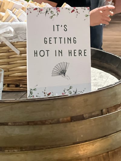 This is a close-up, eye-level, indoor shot of a sign that reads "IT'S GETTING HOT IN HERE" in dark lettering. The sign is white with a floral border at the top and a faded illustration of a hand holding a fan at the bottom, all set against a pale blue background with faint floral accents. The sign is propped inside a dark, circular wooden barrel. To the left of the sign is a woven basket filled with small, individually wrapped items, possibly favors or treats, tied with white ribbons. The background shows a person's hands holding a clear plastic cup. The setting appears to be a venue in Beit Hanan, Israel, possibly a wedding or event. The lighting is natural and somewhat diffused.