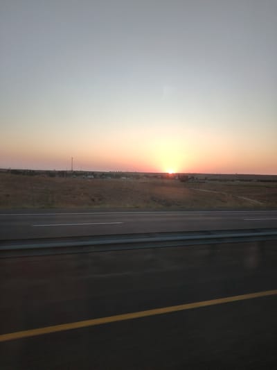 A horizontal, landscape-oriented photograph captures a sunset over a rural area. The sky displays a gradient of colors, transitioning from a pale blue at the top to soft oranges and yellows near the horizon, where the sun is partially visible. The sun's rays cast a warm glow across the scene.
Below the sky, a flat, grassy plain stretches to the horizon. The landscape is dotted with a few trees and small buildings, suggesting a rural settlement. A prominent feature is a tall radio tower to the left of the settlement. The plain appears dry and possibly cultivated.
In the foreground, a paved road runs horizontally across the image. The road has two white lines marking lanes, and a single yellow line runs diagonally across the lower right portion of the frame. A metal guardrail is situated between the road and the grassy plain. The perspective suggests the photograph was taken from a vehicle moving alongside the road. The overall impression is that of a scenic view during dusk in a rural environment, possibly near Samsy, Kazakhstan, given the information provided about the location. There are no people or animals visible in the image. The time of day is clearly sunset.