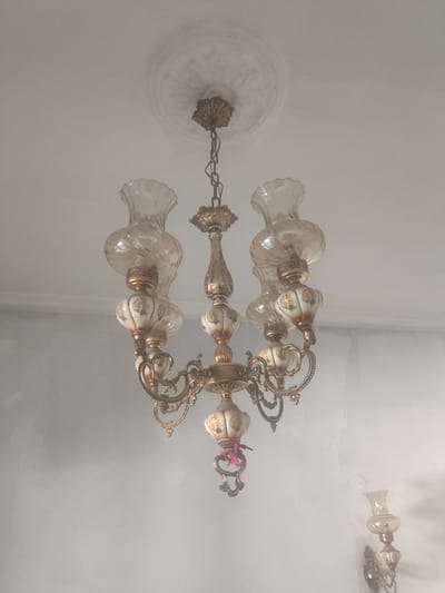 A vintage, ornate chandelier hangs from a ceiling with decorative molding. The chandelier is made of brass with intricate scrolling arms supporting six lamps. Each lamp has a clear, fluted glass shade and a decorative, bulbous ceramic base with floral painted details. The chandelier is illuminated by light bulbs. To the right and lower, a wall sconce with a similar design is visible. The walls are painted a light color. The scene is indoors, likely in a home in Petroupoli, Greece.