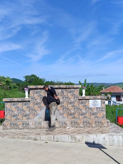 A man in a black shirt and tan pants is standing at a stone fountain on a clear day. He is bent over, appearing to fill a blue water bottle. The fountain is tiled with a mosaic pattern and has white vases with flowers on either side. There is a sign on the fountain with text written in Bulgarian, as well as red buckets attached to the structure. In the background, there is a rural landscape with green hills and a blue sky with wispy clouds. The location is possibly near Medovo, Bulgaria.