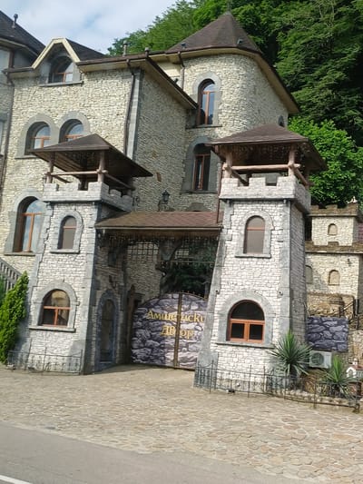 The image shows the entrance to a building with a castle-like facade, located in the city of Kazachiy Brod, Russia. The building is constructed with light-colored stone blocks and features several arched windows, towers, and a large gate with ornamental details. Green trees are visible behind the building. The ground in front of the entrance is paved with cobblestones. The overall architectural style suggests it might be a hotel or a tourist attraction. There are no people visible in the image.