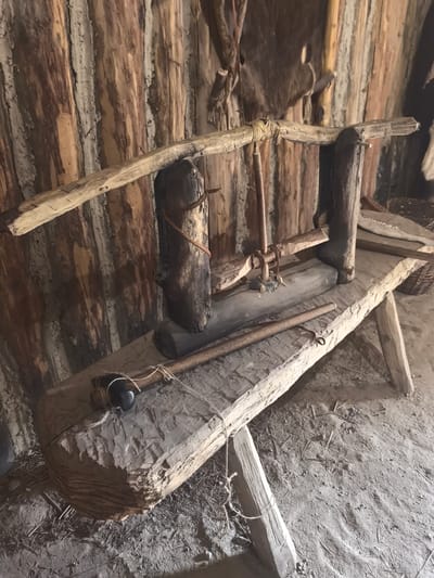 The image depicts an interior scene of what appears to be a historical dwelling, likely a reconstruction or a preserved structure. The walls are constructed from vertical wooden logs, with spaces between them filled with a lighter-colored material, possibly daub or clay. A rough-hewn wooden bench or platform is the central element of the foreground. On this bench, there is a primitive wooden apparatus, possibly for processing materials or as a tool. This apparatus consists of several wooden components, including a beam supported by uprights, a spindle-like element, and a lever. A long, slender wooden stick or rod lies across the bench in front of the main apparatus, with a loop of twine attached to one end, possibly for securing it. The floor is covered in dirt and debris, suggesting a rustic and aged environment. In the background, other wooden elements and possibly animal hides are visible, contributing to the primitive aesthetic. The lighting is natural and somewhat dim, typical of an interior without modern illumination. No people are present in the scene. The image does not provide any information about its location in the city of Камянюцкі сельскі Савет, Belarus.