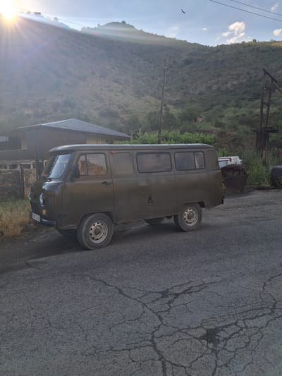 An olive-green UAZ-452 van is parked on a cracked asphalt road in front of a rustic building and a steep, dry, grassy hillside under a bright, sunlit sky with scattered clouds. The van faces left, with its front end slightly angled towards the viewer. Its license plate reads "0580 CK 61". To the left of the van, a low stone fence borders a dry, grassy area. Behind the van, the building appears to be made of corrugated metal roofing and light-colored siding. The background features a dramatic, sun-drenched hillside covered in dry grass and scattered vegetation, with a few trees and power poles visible. The overall scene suggests a rural or semi-rural setting, possibly in Vayk, Armenia, given the landscape and type of vehicle.