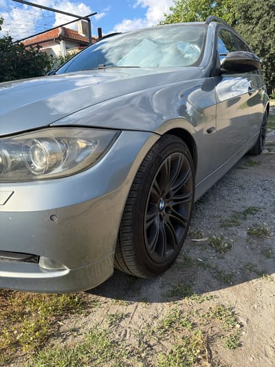 The image shows a close-up, low-angle view of the front passenger side of a silver BMW station wagon. The car is parked on a gravel and dirt surface with some grass and small yellow and white wildflowers growing in patches. In the background, a red-tiled roof of a house is visible, along with some trees and a blue sky with white clouds. The car's headlight, wheel, and part of the fender and door are in focus. The scene appears to be an outdoor setting in a residential area, likely Rusokastro, Bulgaria, given the architectural style of the house and the surrounding vegetation. There are no people or animals visible in the image, and the car is stationary. The car has a sunshade placed on the inside of the windshield.