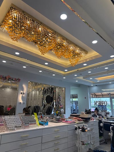The image depicts the brightly lit interior of Chic Nails & Spa, located at 7935 Airport Rd N, Naples, Florida, United States.
The scene focuses on a modern, upscale nail salon. A large, rectangular chandelier, composed of numerous golden and clear crystal-like pendants, dominates the multi-tiered white ceiling, which also features multiple recessed lights.
In the mid-ground, a long, white counter system with numerous drawers, each fitted with gold pull handles, extends horizontally. The countertop is extensively utilized to display hundreds of nail polish bottles in various clear acrylic stands, alongside miscellaneous salon tools, a yellow sign, and other supplies. To the right of the counter, a white, three-tiered rolling utility cart holds brushes and other implements.
Three individuals are present. On the far left, a female client with blonde hair, wearing a pink top, is seated in a salon chair. In the center, behind the counter, a person with dark hair is visible from the back. To the right, near a large window, a female nail technician with dark hair, wearing a dark shirt, is seated at a workstation, actively working with her hands.
The salon's walls are white, with sections adorned with subtle marble-patterned panels and decorative mirrored strips. A prominent, textured circular "C" logo is mounted on a mirrored wall panel in the mid-ground. To its left, a curved, illuminated wall sconce provides accent lighting. On the far right, adjacent to the front window, a decorated Christmas or Easter tree, adorned with pastel eggs and bunny ears, stands as a festive display.
Large front windows reveal an exterior view with palm trees and a parking lot containing vehicles. A reverse "OPEN" sign is visible on the interior side of the glass, indicating the business is operational.