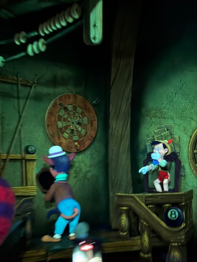 The image captures a scene from the dark ride attraction "Pinocchio's Daring Journey" located in Disneyland Park in Anaheim, California, United States of America. In the foreground, a costumed character resembling Gideon the Cat from the Disney film *Pinocchio* is seen from behind. He is wearing a white hat with a purple band, a teal scarf, and bright blue pants, with his back turned to the viewer. He appears to be interacting with a dark opening in the wall. To the right, Pinocchio, the wooden puppet, is depicted in his iconic outfit of red shorts, a yellow hat with a red feather, and blue gloves. He is shown with his hand over his mouth, a common animated expression. Behind Pinocchio, several framed pictures, possibly a part of the attraction's set design, are visible. A dartboard hangs on the wall between the two characters, with several darts stuck in it. To the left of the dartboard, an eight-ball from a pool game is placed on a wooden shelf. The environment is dimly lit and has a rustic, somewhat spooky atmosphere, characterized by dark green walls and wooden structures.