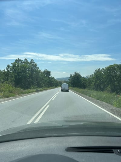 The media file captures a first-person perspective from inside a vehicle, looking out onto an open road in Marino Pole, Bulgaria. The time of day appears to be mid-day, under a bright, clear blue sky adorned with scattered wispy white clouds.

The central element of the scene is a straight asphalt road, receding into the distance. It features double solid white lines in the center, clearly separating lanes of traffic, and a single white line marking the right shoulder. Ahead on the road, a white van is visible, driving in the same direction as the camera. The van's rear license plate, "M 3949 M," is discernible, indicating a Bulgarian registration from the Montana region.

Both sides of the road are bordered by dense green trees and lush vegetation, suggesting a rural or natural setting. Green grass and various wildflowers, including hints of red poppies and white daisies, are visible along the roadside. In the far distance, beyond the trees, the faint outlines of hills or mountains can be seen merging with the horizon.

A portion of the camera's originating vehicle, specifically a dark grey dashboard or pillar, is visible on the right side of the frame, providing context for the view from inside a car. The overall impression is one of a scenic and peaceful drive through a verdant landscape in rural Bulgaria on a clear, sunny day.