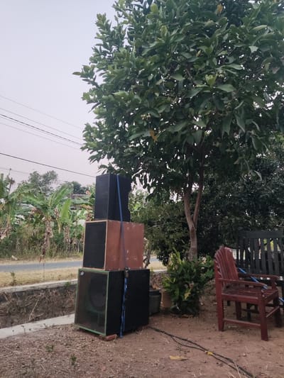 In Ngentrong, Indonesia, the image depicts an outdoor scene with a stack of four speakers in the foreground. The speakers are arranged vertically, with the bottom speaker being the largest and having a green grille. Above it sits a speaker with a brown wooden enclosure and a black grille, followed by another speaker with a black grille and a blue strap. The top speaker is entirely black. To the right of the speakers, there is a red wooden chair and a black wooden bench. Behind the speakers, a large tree with dense green foliage dominates the right side of the frame. In the background, there are banana trees, other vegetation, a road, and power lines stretching across the sky. The ground is covered with dry leaves and dirt.