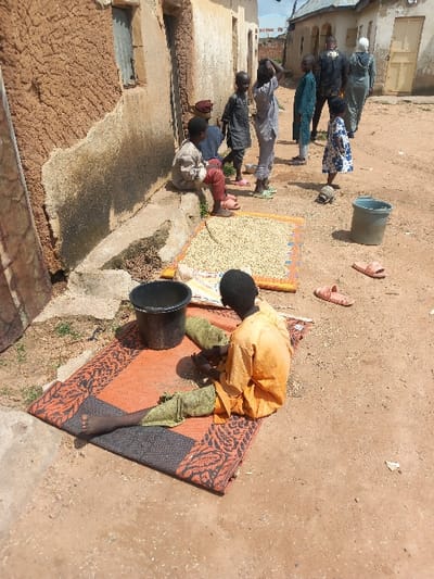 The image depicts an unpaved street scene in Zangon Aya, Nigeria, under daylight. On the left, a weathered building with a mud-plastered facade and concrete base features a window with louvered shutters. In the foreground, an individual wearing an orange long-sleeved garment and green lower wraps sits on a black and orange patterned mat, facing away from the camera, engaged in handling light-colored grains. A black bucket rests beside them on the mat. To their right, a larger patterned mat is spread on the dirt ground, covered with a significant quantity of light-colored, granular material. A gray bucket and two pink slippers are situated near this mat. Along the base of the building on the left, a child in a light-colored top and red pants sits on a concrete ledge. In the midground, a group of five children are present: one in a white and blue patterned dress, another in a light-colored outfit with hands raised to their head, and three others standing. Further down the street in the background, two adults are visible; one male in a dark patterned shirt and one female in a light-colored abaya.