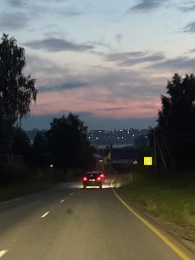 The image depicts a road scene at dusk, likely in the rural settlement of Gavrilovka, Russia. The sky is a gradient of blue, gray, and pink clouds. On the left, a dark silhouette of a birch tree dominates the foreground. A car, with its brake lights illuminated, is driving away from the viewer down the asphalt road, which has white lane markings. To the right of the road, a yellow speed limit sign displaying "40" is visible. In the background, distant lights suggest a populated area. The overall lighting indicates low visibility due to the time of day.