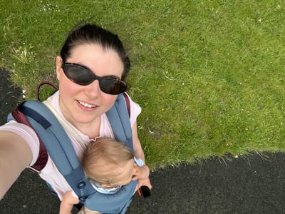 This is a high-angle, selfie-style photograph taken outdoors. A woman wearing dark-rimmed sunglasses is in the foreground, looking at the camera and smiling. She has dark hair pulled back. She is wearing a light pink t-shirt and a blue and burgundy baby carrier. A baby is visible in the carrier, with its head facing away from the camera and its hand near its mouth. The background consists of a grassy area with some small white flowers, bordered by a dark, textured surface, possibly a path or playground surfacing. The scene appears to be in a park or similar outdoor recreational area, common in cities such as Wakefield, United Kingdom.