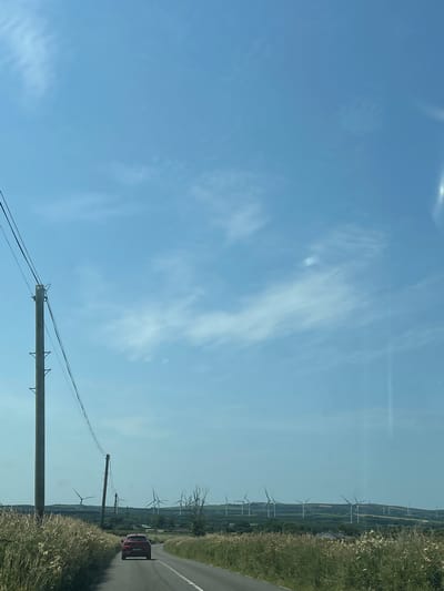 The image depicts a rural roadside scene in Glencullare South, Ireland, under a bright blue sky with scattered wispy clouds. A red car is visible driving away from the camera on an asphalt road that curves to the right. To the left of the road, tall, dry grass and some white wildflowers line the shoulder. A wooden utility pole with multiple wires stretches upwards on the left side of the frame. In the distance, a rolling landscape is dotted with numerous wind turbines, characteristic of the area. The overall environment is open and natural, with a focus on the road, the car, and the distant wind farm.