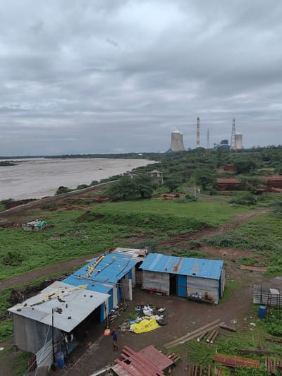 An overcast day in Sudgaon, India, presents a panoramic view of a wide, muddy river to the left, bordered by a concrete embankment. Beyond the river, a lush green landscape stretches towards a distant industrial complex with cooling towers and smokestacks. In the foreground, a collection of makeshift structures with corrugated metal roofs, some painted blue, are situated on a dirt path. Various debris, including yellow tarps, tires, and building materials, are scattered around these dwellings. A single individual, wearing blue clothing, walks near one of the structures. To the right, the ground rises, revealing more green vegetation and scattered trees.