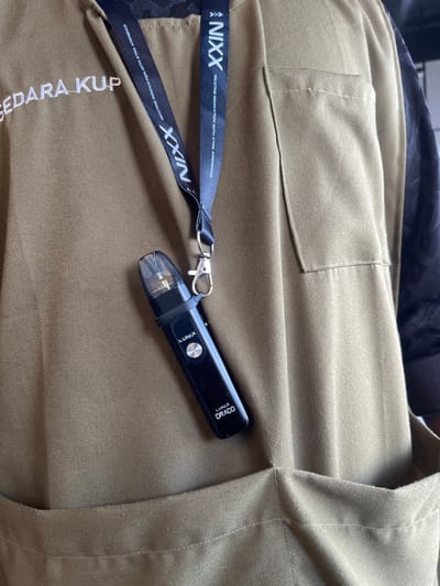 The image shows a close-up of a person's torso, wearing a brown garment with embroidered text on the left side that reads "SEDARA KUP". A black lanyard with white text "NIXX NICOTINE INNOVATION WITH XTRA XPERIENCE" is visible around the neck and draped over the garment. A black vape device, branded "LUNIX DRACO", is attached to the lanyard and hangs near the chest area. The garment has a breast pocket on the right side. The scene is indoors, with the background suggesting a neutral setting. There is no visual information to confirm the location as Bireuenmeunasahdayah, Indonesia.