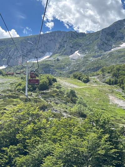 This is a photograph taken from a ski lift in Motički gaj, Montenegro. The ski lift cars, red with yellow seats, are suspended from cables against a backdrop of a rugged, green mountain range. Patches of snow are visible on the mountainside, indicating a high altitude or a lingering winter season. The sky is blue with scattered white clouds. The foreground is filled with lush green trees. The overall impression is a scenic, mountainous landscape, likely captured on a clear day.