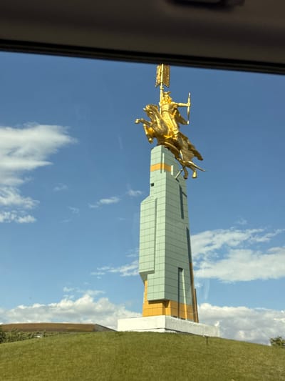 A golden sculpture of a person on horseback is displayed atop a tall, modern building. The building has a pale green facade with accents of orange and white. The sculpture appears to be in motion, with the horse's legs extended and the rider holding a staff. The sky is a clear blue with scattered white clouds. To the right of the building, a grassy hill slopes upwards. The image is taken from a slightly low angle, looking up at the sculpture and building. The scene appears to be outdoors on a sunny day. The overall impression is one of grandeur and achievement, possibly representing a historical or heroic figure. The image is oriented sideways.