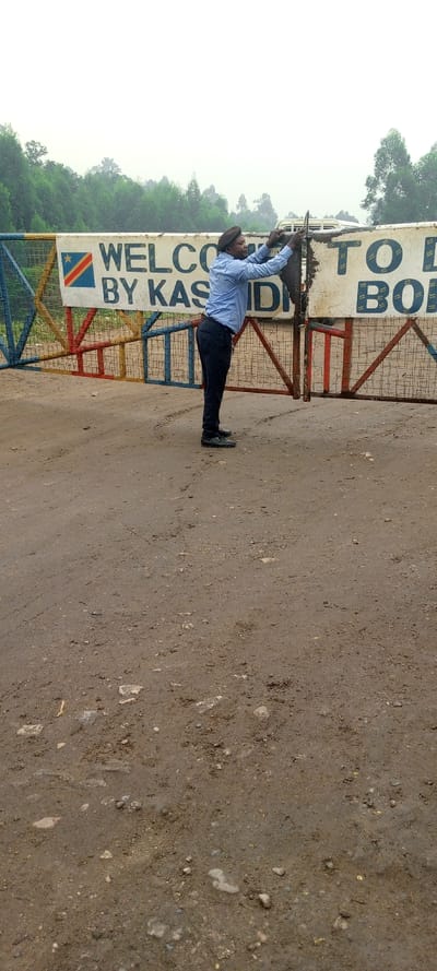 The image shows a man in a blue shirt and dark pants standing at the entrance to Kasindi, a town in North Kivu province of the Democratic Republic of the Congo. The man is opening a metal gate, with a sign behind him that reads “WELCOME TO KASINDI” and features the flag of the Democratic Republic of Congo. The man is wearing a black hat and appears to be in his mid-twenties. The gate is a simple metal gate with a few horizontal bars and some vertical bars. The gate is painted in a variety of colors, including red, blue, yellow, and white. The sign is made of a white metal sheet with black writing and is attached to the gate with screws. The ground is dirt and gravel, and the image is taken from a slight angle to the left of the man. The man is standing in front of the sign, looking to the right, and appearing to be in the process of opening the gate to allow vehicles or pedestrians to enter the town. There are trees in the background, on either side of the gate. The sky is overcast.