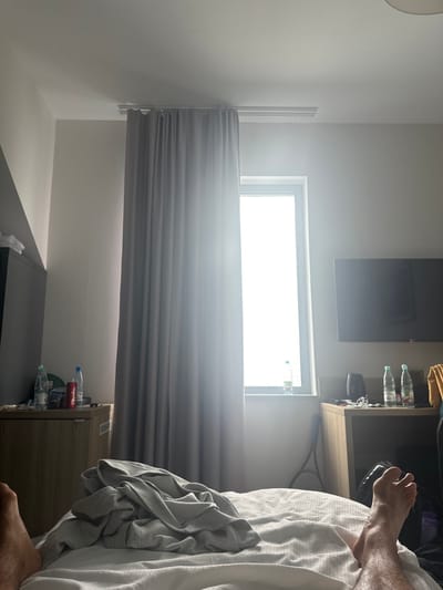 A first-person perspective reveals the interior of a room located in Giebułtów, Poland. The lower legs and bare feet of an individual are visible in the foreground, positioned on a white bedspread. A rumpled grey garment lies on the bed. Directly ahead, a tall, narrow window with a white frame is the dominant light source, emitting strong, overexposed natural light, partially obscured by closed grey curtains.
To the left of the window, a light-colored wooden cabinet or bedside table holds three plastic water bottles and one red soda can. A dark, angled wall or furniture piece is visible in the upper-left background.
To the right of the window, a dark flat-screen television is mounted high on a light-colored wall. Below the television, a matching light-colored wooden table or desk supports two plastic water bottles, a bundle of white coiled cables, a dark electronic device, and a crumpled yellow fabric item. A black tennis racket is leaning against the wall or furniture on the floor near this table. The room's ceiling is white, with a white track system for the curtains visible above the window.
