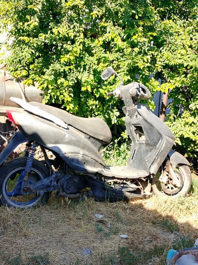 A black scooter is parked on a dry, grassy area with some green bushes and trees in the background. A large metal pipe or tank is partially visible to the left of the scooter. The scooter appears old and weathered, with some parts missing or damaged. The scene is outdoors, under daylight conditions, with shadows cast by the vegetation and the scooter. The location is identified as Pryvilne, Ukraine.