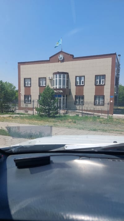 The image shows a two-story building with a beige and brown facade, located in the city of Shakhtinsk, Kazakhstan. A flag of Kazakhstan flies from the roof. The building has several windows, an air conditioning unit, and an entrance with a rounded glass facade. Above the entrance, the word "ПРАТУРА" (PRATURA) is visible in dark letters. A black metal fence surrounds the property, and a small pine tree is planted in front of the entrance. The scene is viewed from inside a vehicle, with the dashboard and windshield wipers visible in the foreground. The sky is clear and blue.