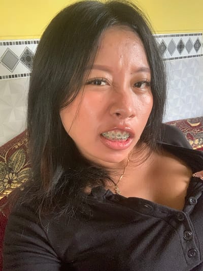 A woman with black hair and braces is looking into the camera. She is wearing a black shirt with buttons. The background consists of a yellow wall and a tiled wall. The scene appears to be indoors, likely in a residential setting in Jajag, Indonesia, as suggested by the style of the walls.